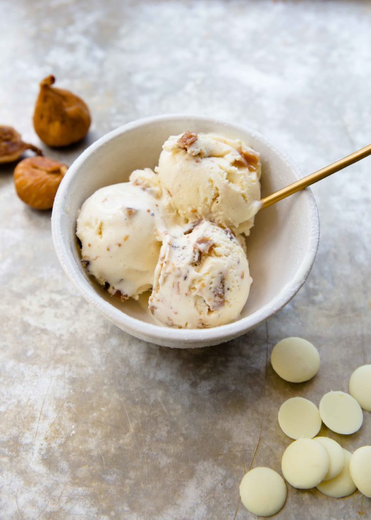 Three scoops of white chocolate fig ice cream in a bowl with white chocolate feves and golden figs.