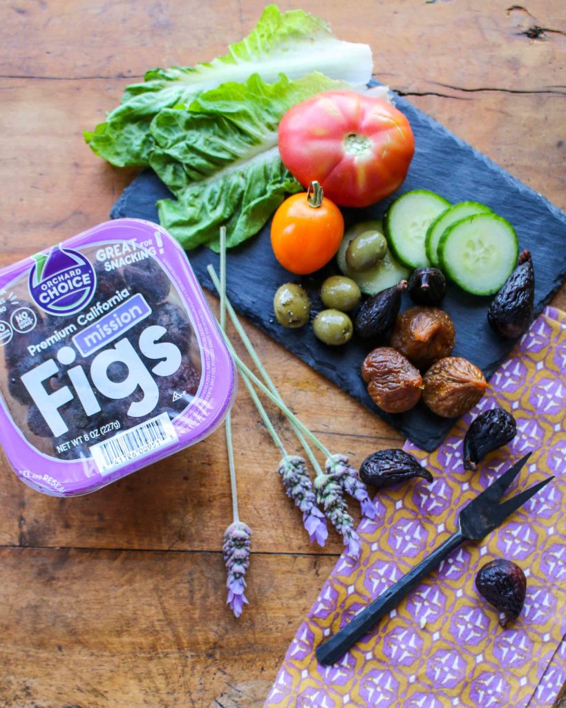 Package of Orchard Choice California Mission Dried Figs, full of plant based fiber, next to lavender sprigs and a tile with olives, tomatoes, and cucumbers on top.