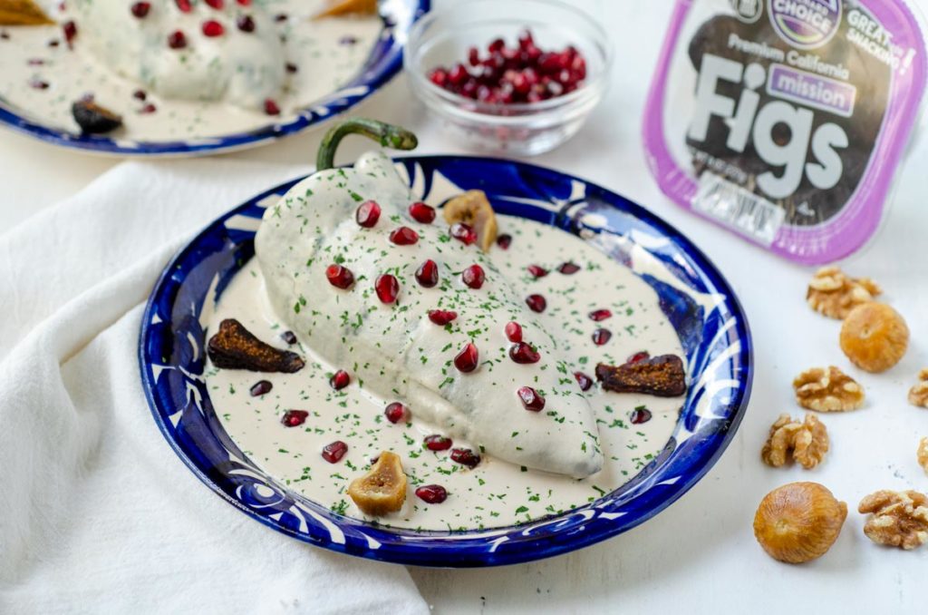 Fig, fruit and nut stuffed poblano peppers covered in a creamy walnut sauce with dried figs around the blue plate and a dish of pomegranate seeds and walnuts in the background with a package of Orchard Choice Mission Figs.