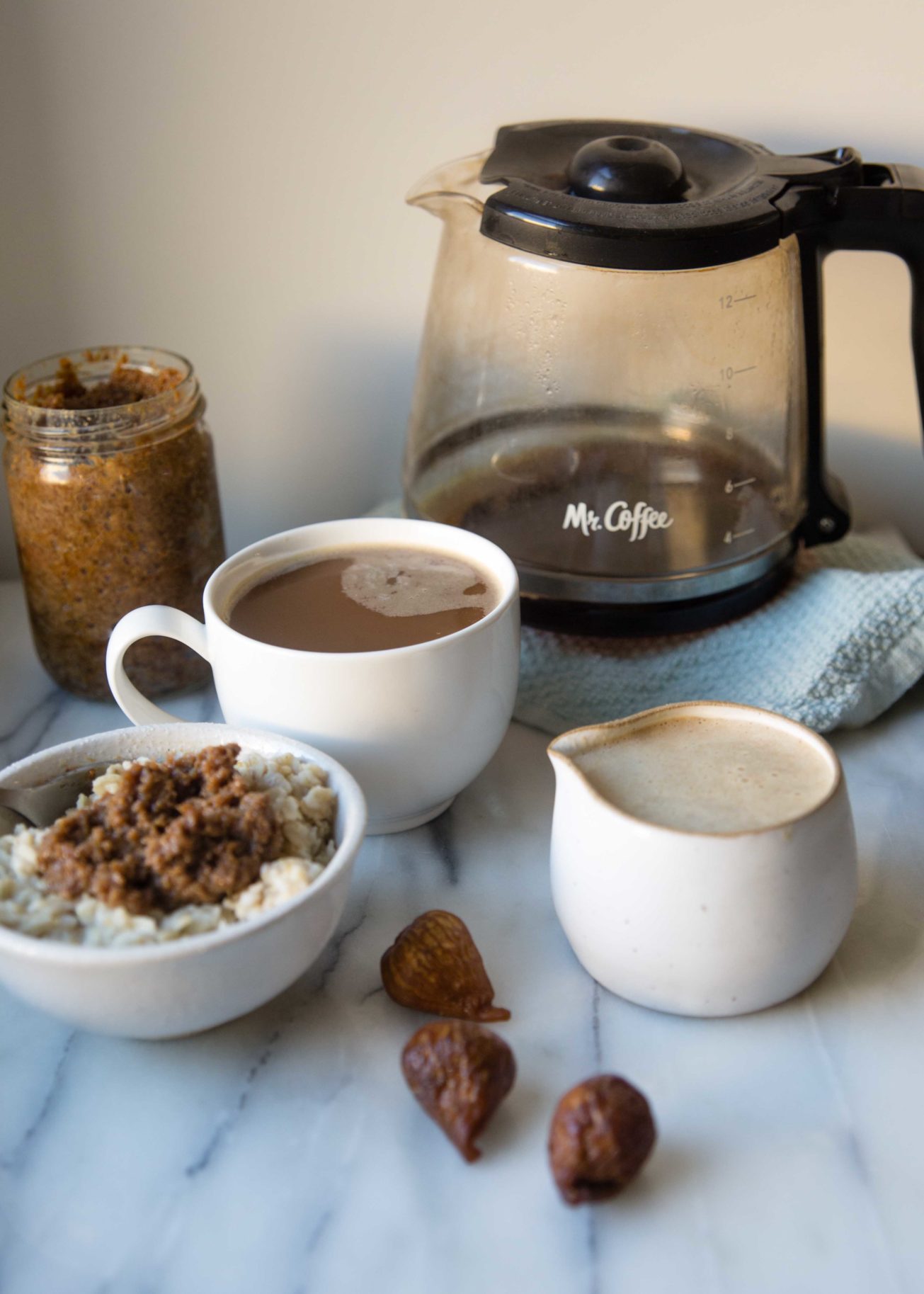 Fig holiday coffee creamer in a creamer receptacle, a coffee pot, coffee mug, bowl of oatmeal topped with Fig Milk Jam, next to a jar of Fig Milk Jam, by Golden Dried Figs on a marble counter.