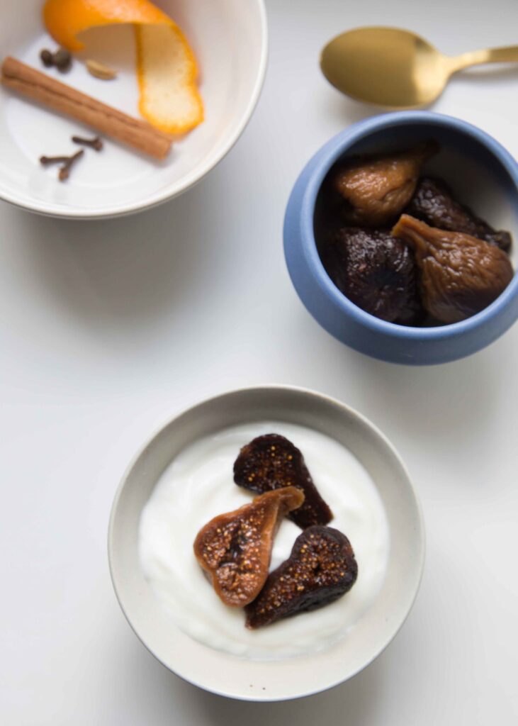 Bowl of stewed figs next to a bowl of halved stewed figs on yogurt with a bowl in the corner that shows orange zest, cinnamon, and cloves.