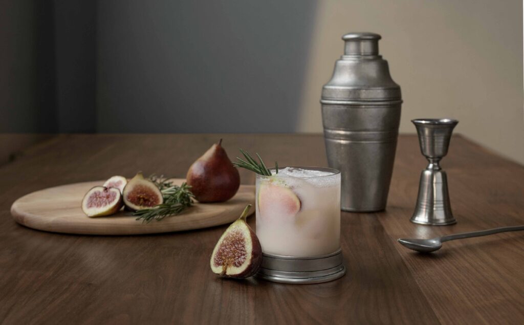 Wooden table with slices of fresh figs a tumbler of milky drink and a stainless steel shaker and measuring spoon nearby