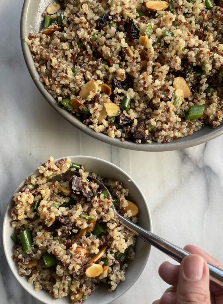 Quinoa Salad with Figs in a grey bowl with a small bowl in front of it with a hand on a spoon, all placed on a marble countertop